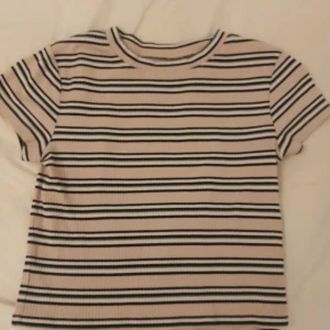 Randig ribbad topp från H&M - Kortärmad topp från H&M i ribbad bomull med svarta, vita och beige ränder. Modellen är klassisk med rund hals och normal passform. Tillverkad i ekologisk bomull, perfekt för en chill och stilren look.