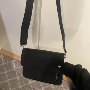 Svart axelväska med bred rem - Säljer en stilren svart axelväska med bred axelrem och guldiga metalldetaljer. Väskan har ett minimalistiskt, fyrkantigt snitt och flera fack inuti för att hålla ordning på dina grejer. Använd fåtal gånger, hör av dig vid funderingar!❣️
