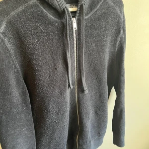 Svart hoodie från Marc O'Polo - Svart hoodie från Marc O'Polo i ekologisk bomull. Tröjan har dragkedja framtill, huva med snörning och långa ärmar. Materialet är stickat och känns mjukt mot huden. Perfekt för dig som gillar enkel och stilren design.