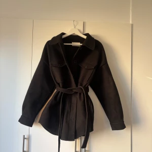 Svart kappa från NLY Trend - Stilren svart overshirt från NLY Trend med bälte i midjan och klassisk krage. Jackan har knappar framtill, två bröstfickor och långa ärmar med knappdetalj vid ärmslut. Perfekt för lager-på-lager och snygg till både jeans och kjol.