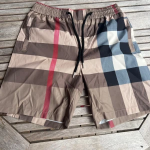 Burberry rutiga shorts med snörning - Snygga Burberry shorts med klassiskt rutigt mönster i beige, brunt, svart, blått och rött. De har elastisk midja med svart snörning och praktiska fickor. Perfekta för sommaren och har en loose passform som ger skön vibe.