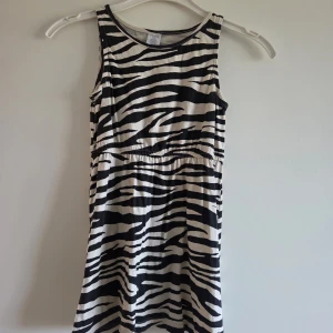 Zebramönstrad klänning från Lindex, stl 110/116 - Snygg zebramönstrad klänning från Lindex i svart och vit. Modellen är ärmlös med rund hals och har en elastisk söm i midjan för skön passform. Tillverkad i 100% bomull som känns mjuk mot huden. Aldrig använd. Har endast hängt i garderoben. 