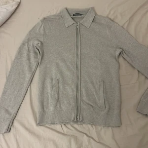 Grå cardigan jacka från Dressmann - Snygg ljusgrå cardigan jacka från Dressmann Premium med dragkedja framtill och två fickor. Jackan är stickad i mjukt material och har klassisk krage samt ribbade muddar vid ärmslut och nederkant. Perfekt för chill dagar och enkel att matcha.