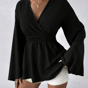 Svart tröja med knyt i ryggen - Svart omlottopp med v-ringning och långa, vida ärmar. Toppen har smockad midja som ger en snygg siluett och knytning i ryggen för extra detalj. Materialet är lätt och perfekt för att skapa en chill och trendig look.