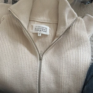 Beige stickad kofta från Maison Margiela - Säljer en beige stickad kofta från Maison Margiela med ribbad struktur och hög krage. Koftan har en snygg dragkedja framtill och långärmade ärmar. Perfekt för dig som gillar minimalistisk och clean stil. Passar s med 