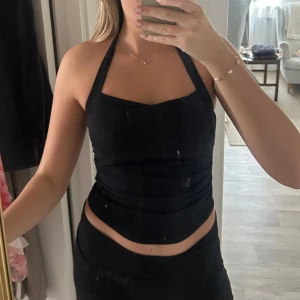 Svart halterneck topp - Snygg svart halterneck topp med croppad passform och knyt i nacken. Toppen har en stilren, slät design och är ärmlös med öppen rygg. Perfekt för dig som gillar minimalistisk stil och vill ha något som känns både trendigt och bekvämt.