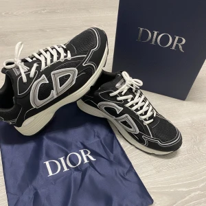 Dior skor  - Säljer ett par svarta Dior sneakers med vita kontrasterande detaljer och stora CD-loggor på sidorna. Skorna har vit snörning och en chunky vit sula. Materialet är en mix av mesh och syntet, vilket ger en sportig och modern vibe. Perfekta för dig som gillar exklusiv streetstyle.