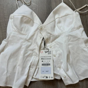 Vit korsett-topp med knappar XS - Snygg vit korsett-topp med smala axelband och knappar framtill. Toppen har en figurnära passform och markerade kupor som ger en cool siluett. Perfekt att styla med jeans eller kjol för en trendig look.