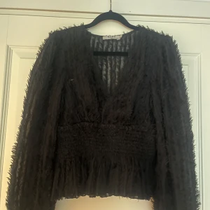 Svart fluffig blus från NA-KD - Säljer en svart blus från NA-KD i storlek 34. Blusen har en fluffig, texturerad yta och djup v-ringning. Midjan är smockad för en snygg siluett och ärmarna är långa. Perfekt för dig som gillar statement-plagg med cool struktur.
