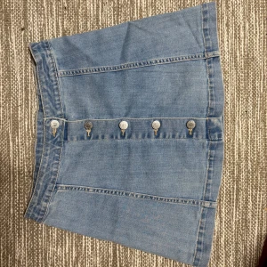 Blå jeanskjol med knappar från Cubus stl.158 - Snygg ljusblå jeanskjol från Cubus med raka linjer och fem silverfärgade knappar framtill. Stl.158 (ställbar midja)Kjolen är kort och har klassisk denimlook med synliga sömmar. Materialet är 80% bomull, 18% polyester och 2% elastan för lite stretch. Perfekt för dig som gillar enkel och cool stil.