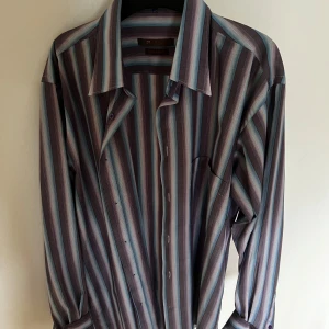 Randig skjorta från Brice i blått och lila - Snygg skjorta från Brice med vertikala ränder i blått, lila och beige. Modellen har klassisk krage, lång ärm och knappar framtill. Passformen är slim och materialet känns som bomull. Perfekt för dig som gillar färg och mönster.