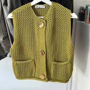 Stickad grön väst från Zara - Säljer en chunky stickad väst från Zara i en cool olivgrön färg. Västen har grov struktur, två fickor fram och stora, snygga knappar. Perfekt att slänga över en t-shirt för en chill look. Passformen är normal och materialet känns mjukt och skönt.