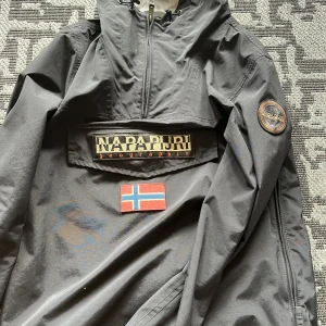 Svart vindjacka från Napapijri - Svart vindjacka från Napapijri med halv dragkedja, huva med snörning och stor logga på bröstet samt norsk flagga.  Perfekt inför hösten! Nypris 2200kr