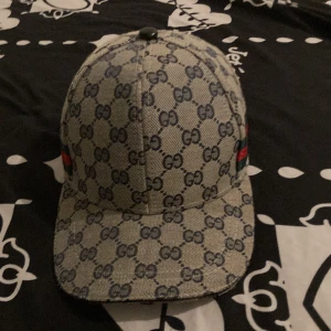 Gucci keps med GG-mönster och läderdetalj - Snygg keps från Gucci i beige och marinblått GG-mönster. Klassisk böjd skärm, justerbar rem i svart läder bak och ikoniska gröna/röda ränder på sidan. Tillverkad i bomullscanvas med exklusiv känsla och tydliga logodetaljer invändigt.