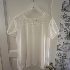 Vit blus med spets från Soyaconcept - Vit blus från Soyaconcept med korta ärmar och snygga spetsdetaljer upptill och på ärmarna. Blusen har rund hals och en liten knapp i nacken. Tillverkad i mjuk polyester som känns lätt och skön mot huden. Perfekt för dig som gillar stilrena plagg. 