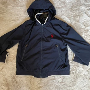 Mörkblå vindjacka från Polo Ralph Lauren - Hej! Jag säljer nu en marinblå vindjacka från Polo Ralph Lauren. Jackan är i mycket gott skick utan defekter. Jag som är 183cm var växt ur jackan och därför säljer jag den. Min uppskattning är att den borde passa någon som är omkring 175cm beroende på vilken passform man vill ha. Priset är inte hugget i sten, vänligen kontakta mig vid minsta fundering! Efter ett köp fraktas tröjan inom 24h🤩