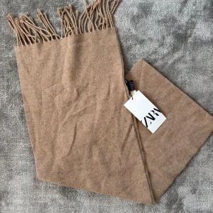 Beige halsduk från Zara - Snygg beige halsduk från Zara med klassiska fransar i kanterna. Den är mjuk och har en enkel, stilren design som gör den lätt att matcha med olika outfits. Perfekt för att hålla dig varm under kyliga dagar. Hör av dig vid frågor och funderingar!!