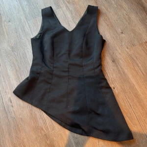 Svart asymmetrisk topp Gina Tricot - Svart topp från Gina Tricot i storlek 36 med snygg asymmetrisk nederdel och v-ringad hals. Toppen  är ärmlös och har en diskret dragkedja i sidan för enkel påklädning. Materialet är mjukt och följsamt, perfekt för dig som gillar stilrena och moderna plagg.