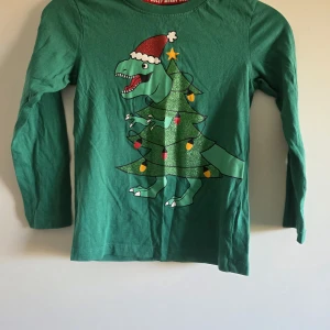 Grön långärmad tröja med julmotiv - Rolig grön långärmad tröja i bomull med tryck av en dinosaurie utklädd till julgran, pyntad med glitter, lampor och tomteluva. Perfekt för dig som vill sticka ut på julen! Halsen har ett rött band med texten 'Jolly Merry'.
