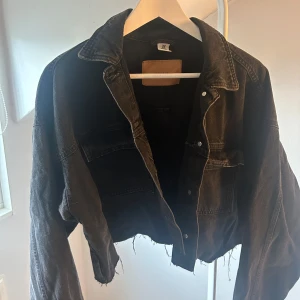 Svart croppad jeansjacka - Säljer en svart croppad jeansjacka från H&M med rå kant nedtill och klassisk krage. Jackan har långa ärmar, två bröstfickor med lock och knappar framtill. Perfekt för dig som gillar en edgy och avslappnad stil.