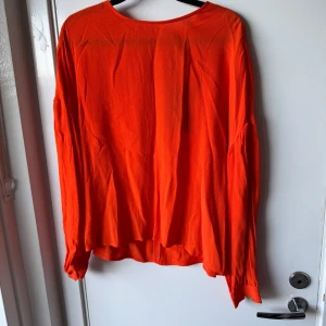 Orange blus från Stradivarius - Snygg orange blus från Stradivarius med rund hals och långa ärmar. Blusen har en lätt struktur i tyget och en liten knapp i nacken. Perfekt för dig som vill sticka ut med färg! Materialet känns mjukt och luftigt mot huden.