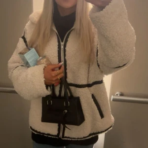 Vit teddyjacka med svarta detaljer - Säljer en fluffig vit teddyjacka med svarta kontrastdetaljer och dragkedja framtill. Jackan har långärmade, oversized ärmar och två fickor med svarta kanter. Perfekt för dig som vill ha en mysig och trendig look under kyliga dagar. Använts ett fåtal gånger under hösten.