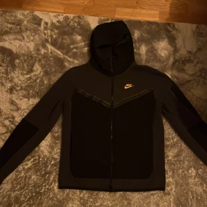 Svart grå Nike tech kofta old season - Nike tech kofta i svart och grå med guldigt logotryck på bröstet. Jackan har hel dragkedja framtill, huva och långärmade ärmar. Snygga detaljer med svarta paneler och en diskret ficka med dragkedja på ärmen. Passar både S-M