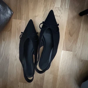 Svarta ballerinaskor med rosett - Snygga svarta ballerinaskor med spetsig tå och liten rosett framtill. Skorna har öppen häl och en elegant, tunn rem runt hälen. Materialet ser ut att vara mocka eller syntet med matt finish. Perfekta för dig som gillar stilrena och trendiga skor. Använda ca 2 ggr sedan köp!💘