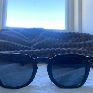 Svarta retroinspirerade solglasögon - Snygga svarta solglasögon med klassisk, lite tjockare båge och runda glas. Bågarna har små silvriga detaljer vid gångjärnen som ger en cool vintagekänsla. Perfekta för dig som vill ha en stilren och tidlös look.