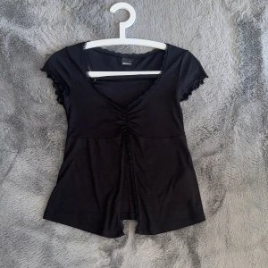 Svart omlottopp från Gina Tricot - Supersnygg svart omlottopp från Gina Tricot med v-ringning och rynkdetalj framtill. Toppen har korta ärmar med volangkant och spets runt halsen. Mjuk och stretchig bomullskvalitet som sitter bekvämt och ger en feminin siluett.