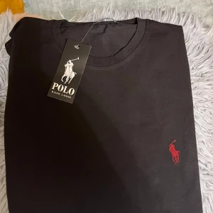 Svart t-shirt från Polo Ralph Lauren - Svart t-shirt från Polo Ralph Lauren i mjuk bomull. Klassisk rund hals och korta ärmar. På bröstet finns den ikoniska röda Polo-loggan broderad. Perfekt basic med stilren design och snygg passform.