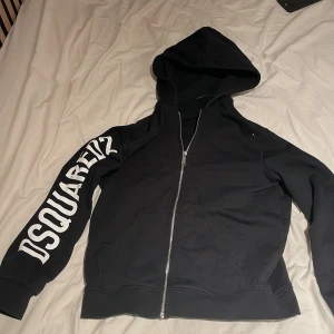 Svart DSQUARED2 hoodie med tryck - Svart hoodie från DSQUARED2 med stor vit logotyp längs ena ärmen och silverfärgad dragkedja framtill. Tröjan har huva, känguruficka och är tillverkad i mjuk bomull. Perfekt för dig som gillar streetwear och vill sticka ut med snyggt tryck.