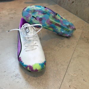 Puma King fotbollsskor i vitt och lila - Säljer ett par Puma King fotbollsskor med coolt färgstänk i lila, blått, grönt och gult. Skorna har vit ovandel med svarta detaljer, snörning och en färgglad sula med dobbar. Insidan är fodrad i lila och hälen har KING-tryck. Perfekt för dig som vill sticka ut på planen!