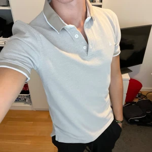 Beige pikétröja från Jack & Jones - Snygg beige pikétröja från Jack & Jones med vit kant på krage och ärmslut. Klassisk polokrage med tre knappar och diskret logga broderad på bröstet. Tröjan är gjord i mjuk bomull och har en normal passform som sitter skönt.