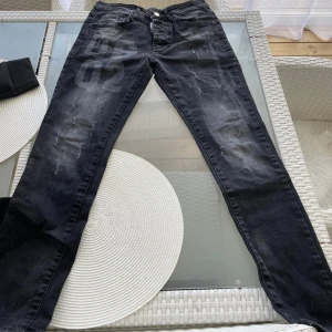 Svarta jeans med DS02-tryck - Svarta jeans från DSQUARED2 med DS02-tryck på vänster lår. Jeansen har en slim passform, klassisk femficksdesign och subtila slitningar för en cool, streetad look. Materialet är jeans och färgen är mörk svart med lite tvättade detaljer.