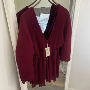 Vinröd plisserad kortklänning från NA-KD - Supersnygg vinröd kortklänning från NA-KD med plisserat tyg och v-ringad hals. Klänningen har långa ärmar och ett flowy, luftigt fall som ger en cool och trendig vibe. Perfekt för dig som vill sticka ut med en djup färg och skön passform.