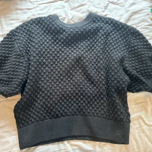 Svart stickad tröja med struktur - Säljer en svart stickad tröja med coolt bubbligt strukturmönster. Tröjan har rund hals och korta ärmar, samt bred ribbad kant nedtill och vid halsen. Perfekt för dig som gillar en unik och chill stil.