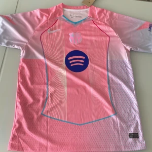 Barça rosa fotbollströja Nike Rikardo 10 - Snygg FC Barcelona fotbollströja i rosa med blå detaljer och tryck. Kortärmad modell med V-ringad krage och tunna vita linjer på framsidan. Spotify-logga och klubbmärke på bröstet, Rikardo 10 och UNHCR-tryck på ryggen. Tillverkad i lätt, ventilerande polyester.