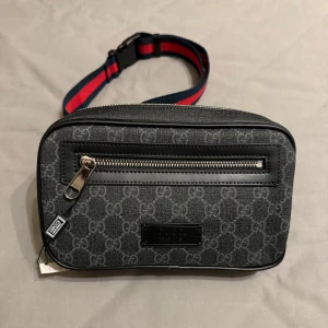 Svart Gg axelväska med monogram - Snygg svart axelväska från Gg med klassiskt GG-monogram i grått. Väskan har en silverfärgad dragkedja framtill och en justerbar rem i marinblått och rött. Materialet är en mix av skinn och polyester, perfekt för att bära det viktigaste.
