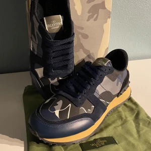 Valentino Garavani Rockrunners - Snygga Rockrunners med mörkblå och grå camouflagemönster, svarta mockadetaljer och beige sula. Skorna har rund tå, mörkblå snörning och feta nitar på hälen. Materialmix av skinn, mocka och textil. Perfekt nu till hösten och vintern. Köpte fel storlek och därför säljer jag. Box, dustbag, kort och extra skosnören följer med. Kom gärna med frågor och tveka inte på att lägga ett bud:)