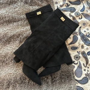 Svarta mockastövlar med spetsig tå - Säljer ett par svarta stövlar i mjuk mocka med spetsig tå och låg klack. Stövlarna har snygga guldfärgade detaljer upptill och dragkedja på insidan för enkel påtagning. Perfekta för dig som vill ha en stilren och edgy look.