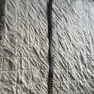 Svart Moschino bälte med gulddetalj - Snyggt svart bälte från Moschino med stor guldfärgad logotyp framtill. Bältet är smalt och har en stilren design som passar till jeans eller klänning. Tillverkat i skinn med en klassisk form och justerbar längd. Perfekt för dig som gillar statement-accessoarer.