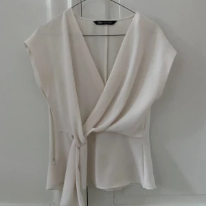 Vit omlottblus från Zara - Snygg vit omlottopp från Zara med knytning i sidan. Toppen har en elegant v-ringning och är ärmlös, vilket gör den perfekt för varmare dagar. Materialet är mjukt och följsamt, vilket ger en skön passform.