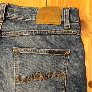Blå jeans från Nudie Jeans - Säljer ett par klassiska blå jeans från Nudie Jeans med snygga slitningar och kontrastsömmar. Jeansen har fem fickor, orange broderi på bakfickorna och en skinnpatch med logga i midjan. Perfekt passform och riktigt skön denimkänsla.