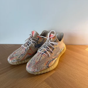 Adidas Yeezy Boost 350 V2 mönstrade sneakers - Säljer ett par Adidas Yeezy Boost 350 V2 med färgstarkt mönster i beige, blått, rött och gult. Skorna är i storleken 44. Skorna har stickad ovandel, snörning och en mjuk, ribbad platt sula med boost-teknologi. Perfekt för dig som gillar unika och trendiga sneakers.