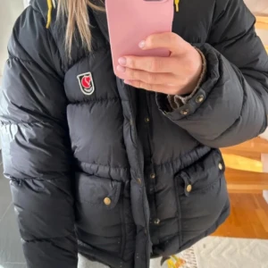 Svart dunjacka från Fjällräven - Snygg svart dunjacka från Fjällräven med gula snören i huvan och klassisk logga på bröstet. Jackan har stora fickor med knappar, dragkedja och tryckknappar framtill.. använd väldigt fåtal gånger