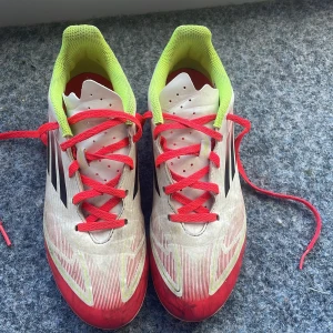 Adidas vita och röda fotbollsskor - Säljer ett par Adidas fotbollsskor med snygg design i vitt, rött och neon-gult. Skorna har röda snören, svarta Adidas-ränder på sidan och en låg profil med platt sula och dobbar för gräsplan. Ovandelen är i syntetmaterial och insidan har mesh för extra ventilation.