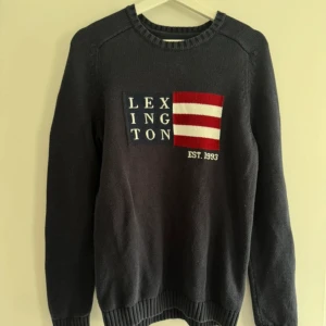 Mörkblå Lexington stickad tröja - Snygg mörkblå stickad tröja från Lexington i storlek S. Tröjan har ett broderat Lexington-flagga tryck med röda och vita ränder på bröstet och texten 'EST. 1993'. Tillverkad i 100% bomull med ribbade muddar och rund hals. Perfekt för dig som gillar klassisk college-stil.. Priset kan också diskuteras!