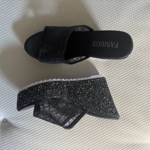 Svarta glittriga sandaler  - Använt enstaka gång. ett par svarta sandaler med glittrig plattform och bred rem i spets. Snyggt mönstrad mesh på ovansidan och öppen tå. Sulan är vit med stjärndetalj undertill.