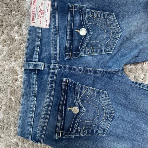 True Religion Joey Low Rise Flare Jeans - Säljer ett par blå True Religion Joey jeans med låg midja och flare-ben. Klassiska kontrastsömmar, stora fickor med lock och silverknappar bak. Jeansen är använda fåtal ggr och från nelly i strl 27, säljs för kommer inte till användning!! 